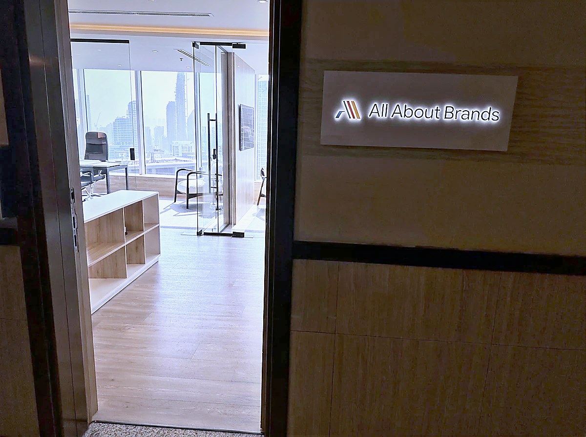 The open door of the All About Brands office in Business Bay, the illuminated company sign on the travertine wall beside it and the Dubai skyline through the windows inside