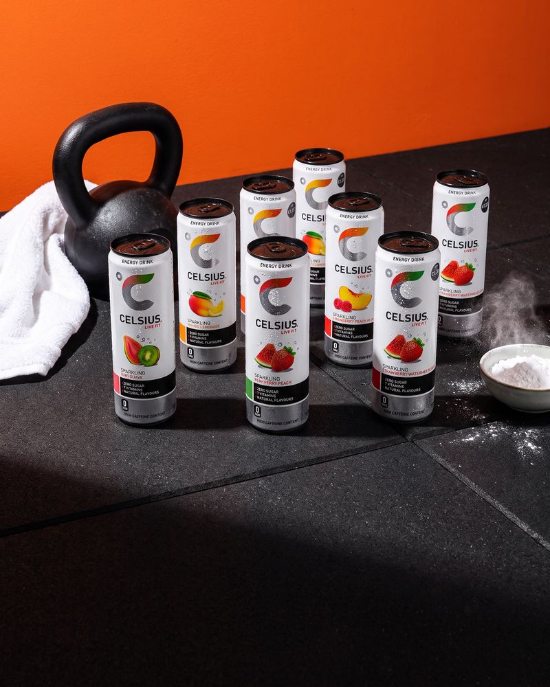 Eight Celsius energy drink cans in Kiwi Guava, Mango Lemonade, Raspberry Peach and Strawberry Watermelon on a black rubber gym floor beside a kettlebell, a towel and a bowl of chalk, against an orange wall.