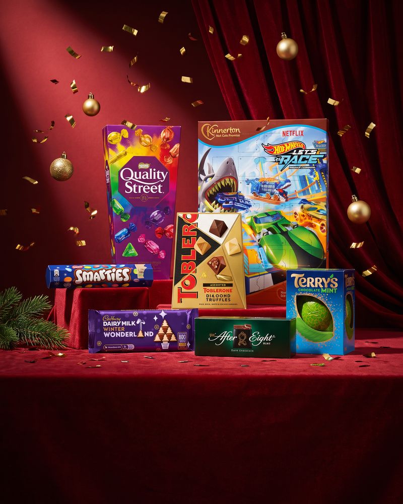 Chocolate gift tins, selection boxes and gold foil-wrapped chocolates arranged on a deep burgundy background.