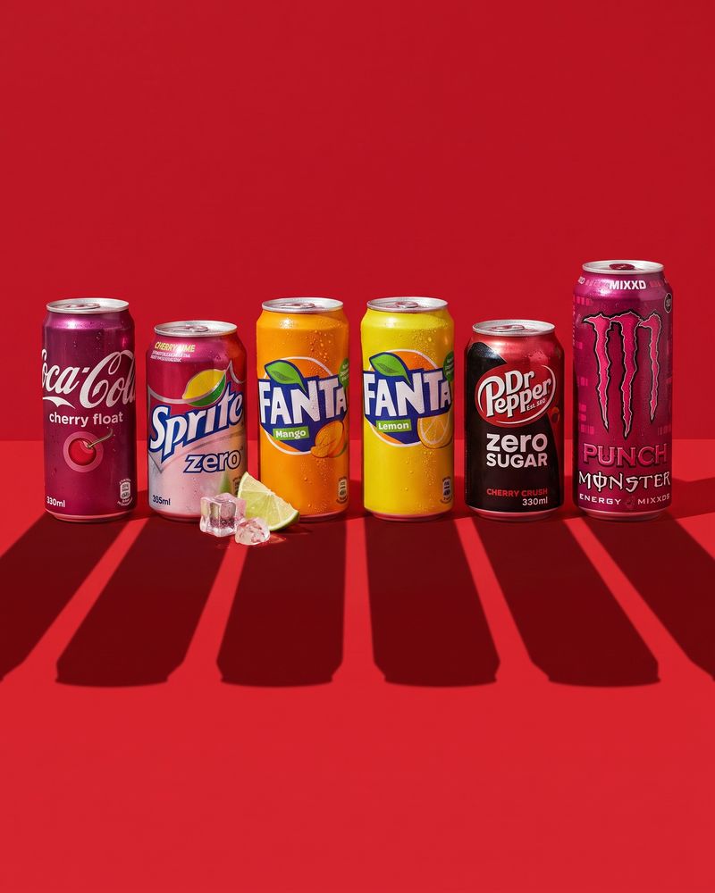 Chilled bottles and cans of cola with ice cubes and citrus slices, on a deep red background.