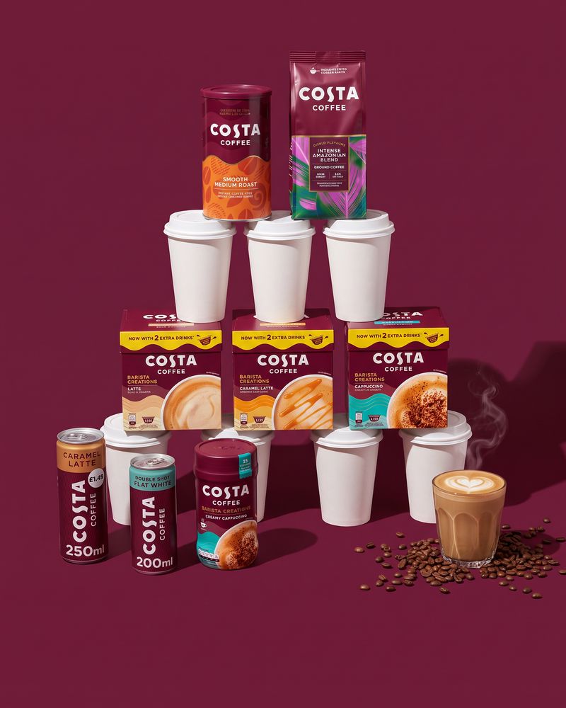 Costa Coffee Dolce Gusto pod boxes, an instant coffee tin, a ground coffee bag, two cans and a Barista Creations tub standing on a pyramid of white takeaway cups beside a glass of latte, on a deep burgundy backdrop