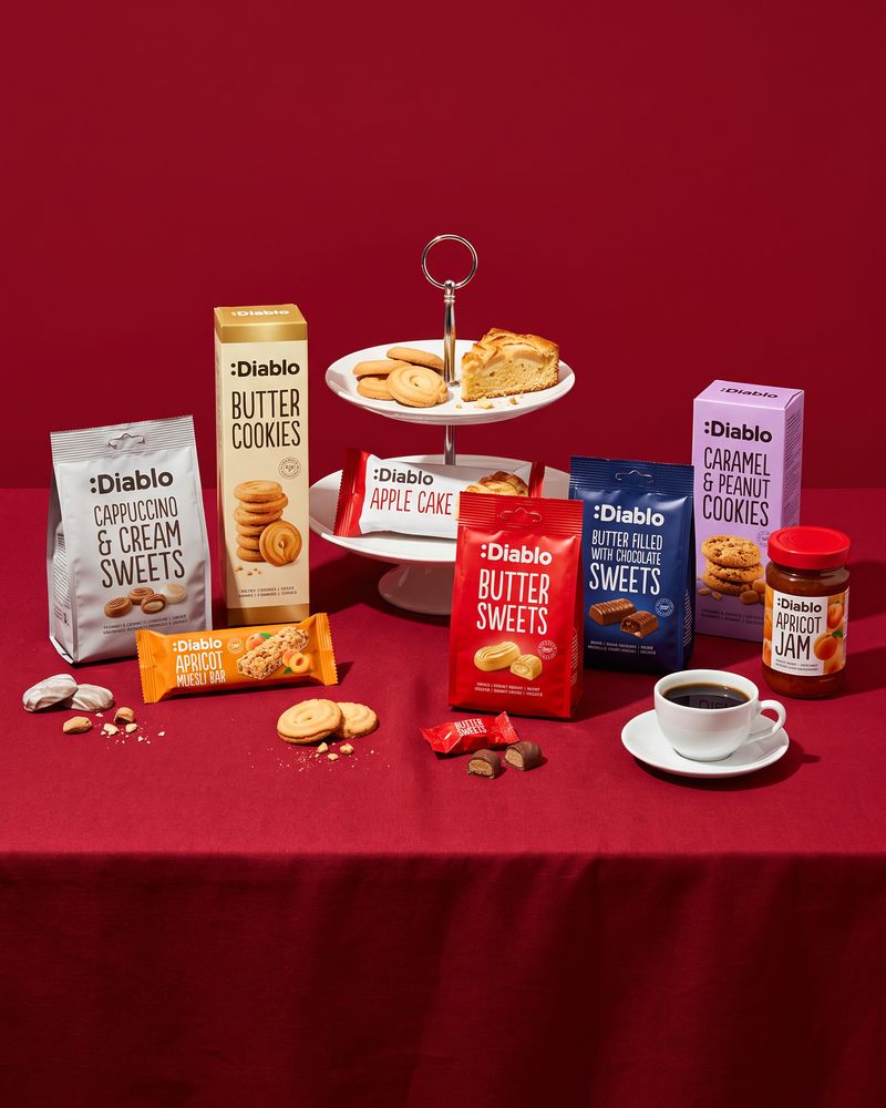 Diablo sugar-free sweets, butter cookies, apple cake, a muesli bar and apricot jam around a white cake stand and a cup of coffee on a deep red tablecloth