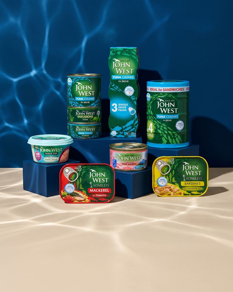 A stack of green John West tuna tins beside a three-pack sleeve and a four-can multipack, with a fridge pot, a pink salmon tin and red mackerel and yellow sardine tins on navy plinths, rippling water light on a deep navy backdrop and a pale sand floor.