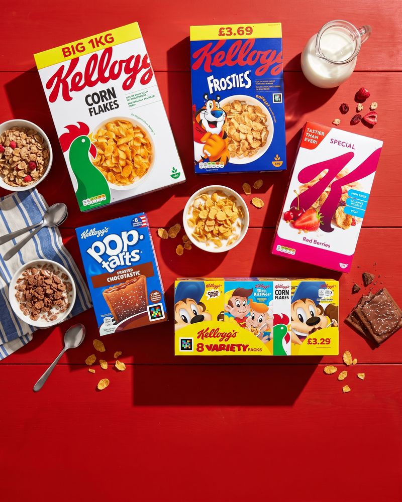 Cereal cartons behind a spill of corn flakes and a splash of milk, on a warm red background.