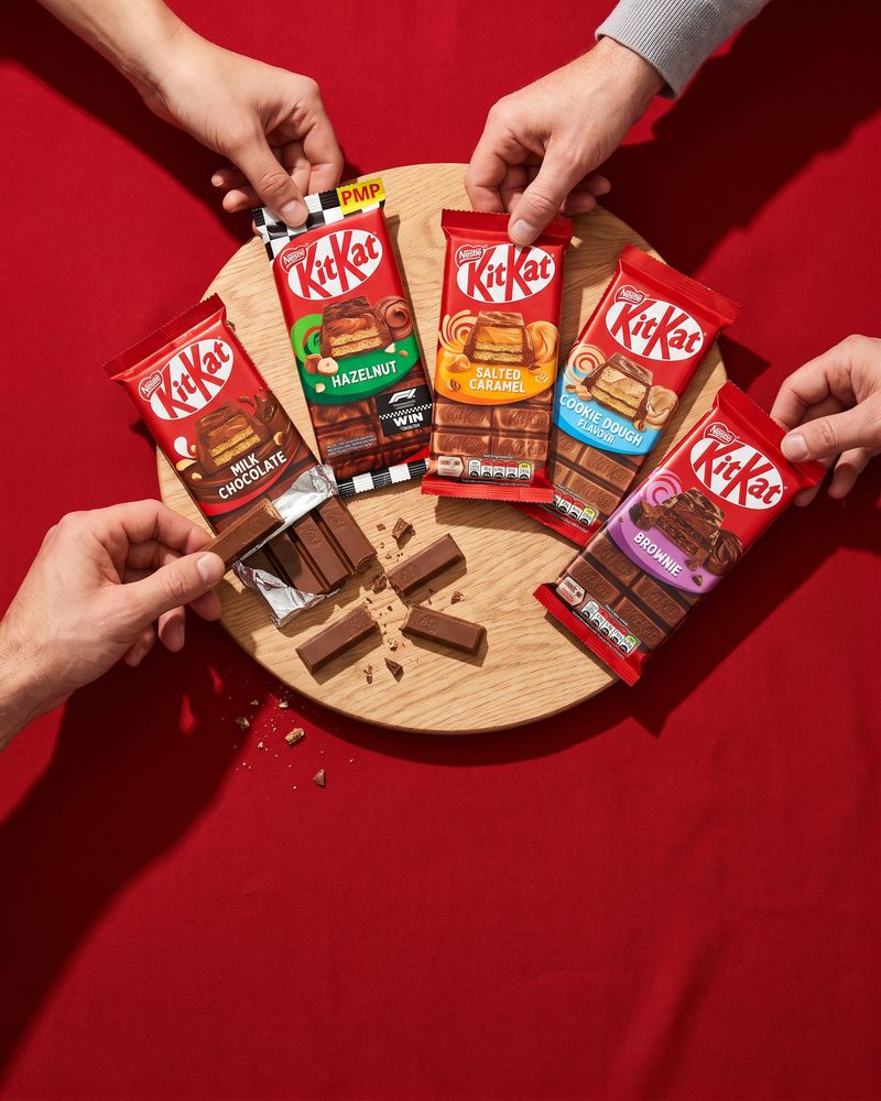KitKat sharing blocks in their red wrappers and flavour packs, with one block snapped open to show the wafer fingers, against a deep KitKat-red backdrop