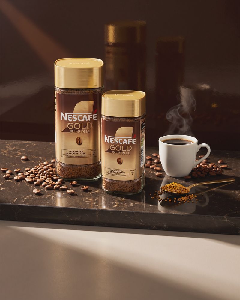 Two Nescafe Gold Blend jars, the 190g and the 95g, standing on a dark stone café counter among loose roasted coffee beans, a white cup of black coffee and a brass spoon of golden granules, against a deep espresso-brown backdrop