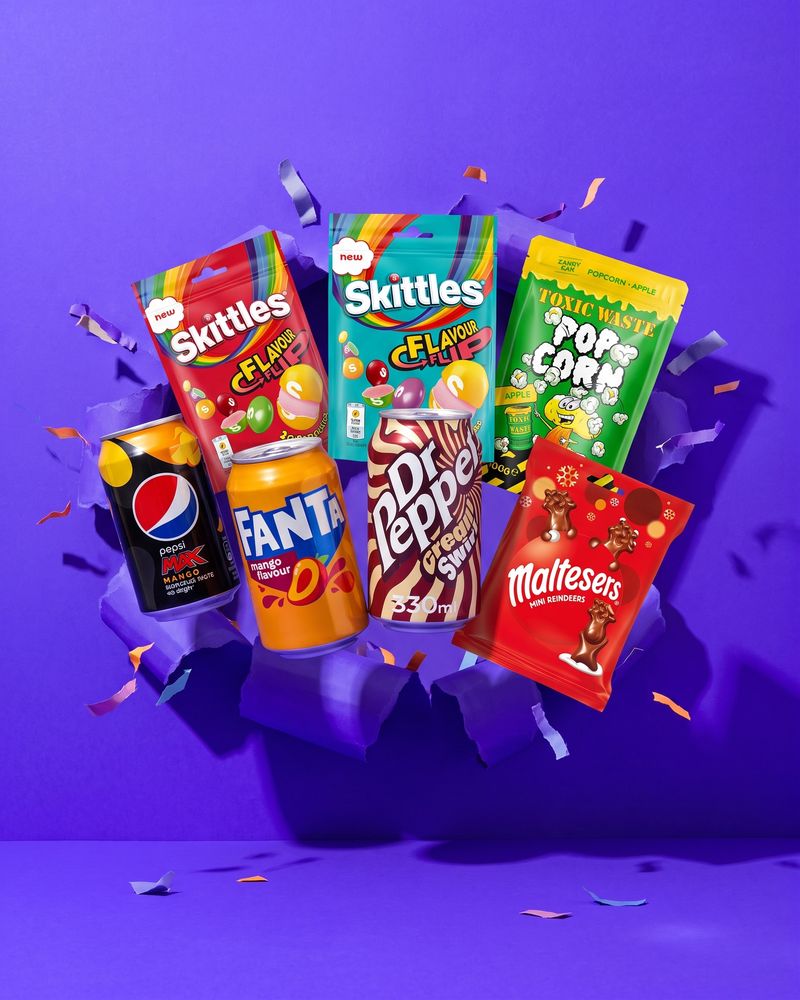An assortment of new grocery and confectionery packets standing upright on an electric violet background.