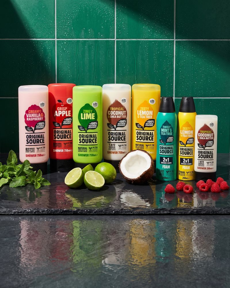 Five Original Source 750ml shower gel bottles, two Foamburst cans and a 250ml bottle standing in a row on a wet black slate shelf among mint sprigs, halved limes, a halved coconut and raspberries, in front of a wall of deep green bathroom tiles