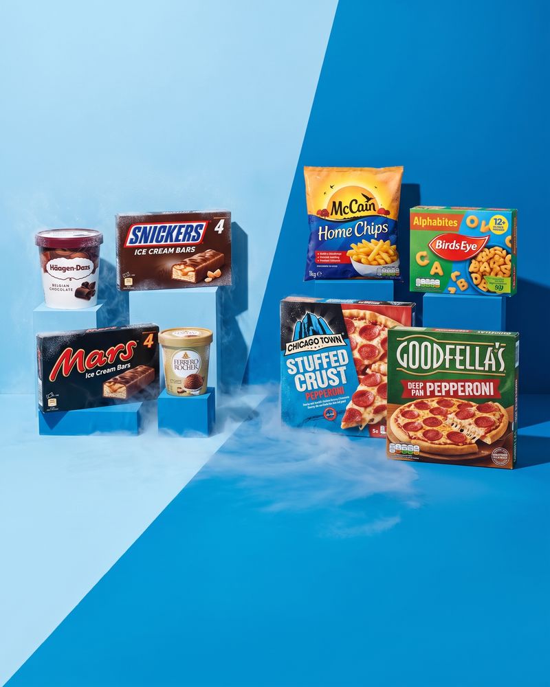 Ice cream tubs and multipack boxes from Häagen-Dazs, Mars, Snickers and Ferrero Rocher on the pale half of a diagonally split blue studio backdrop, with McCain chips, Birds Eye Alphabites and two pepperoni pizza boxes on the dark half, dry ice drifting at floor level.