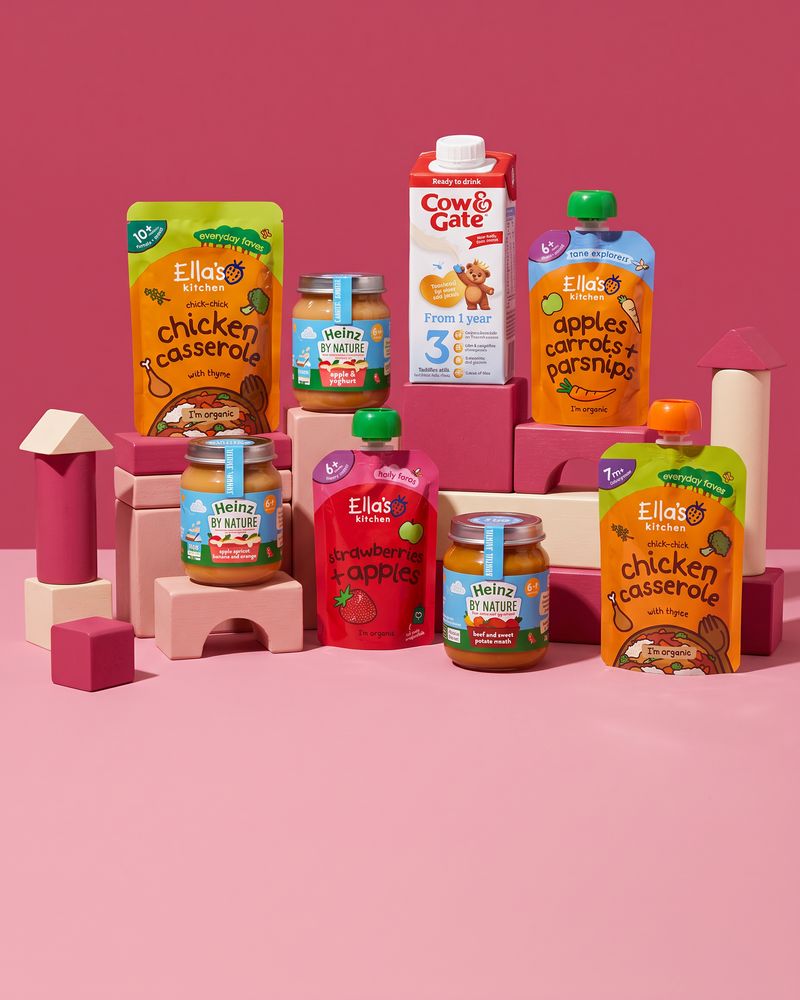 Ella's Kitchen pouches, Heinz By Nature jars and a Cow & Gate growing-up milk carton standing on raspberry-pink and cream wooden toy blocks against a pink backdrop.