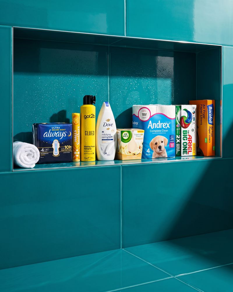 Always Ultra Secure Night pads, Halls Soothers, got2b Glued hairspray, Dove caring bath, an Air Wick vanilla candle, Andrex Complete Clean, Ariel Big One pods and Bacofoil air fryer liners lined up in a glossy teal-tiled bathroom niche beside a rolled white towel.