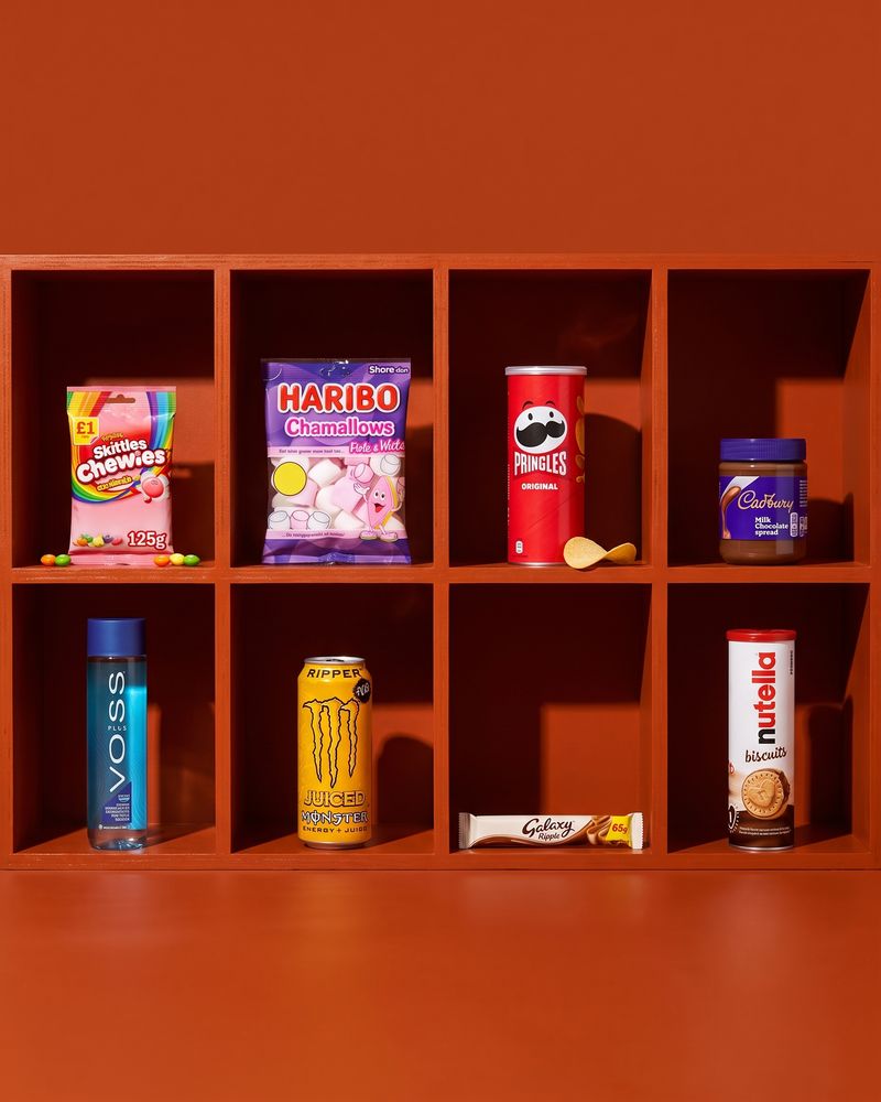 Eight everyday lines each in its own burnt-orange pigeonhole — a Skittles bag, Haribo Chamallows, a Pringles tube, a Cadbury chocolate spread jar, a Voss water bottle, a Monster Ripper can, a Galaxy Ripple bar and a Nutella Biscuits tube.
