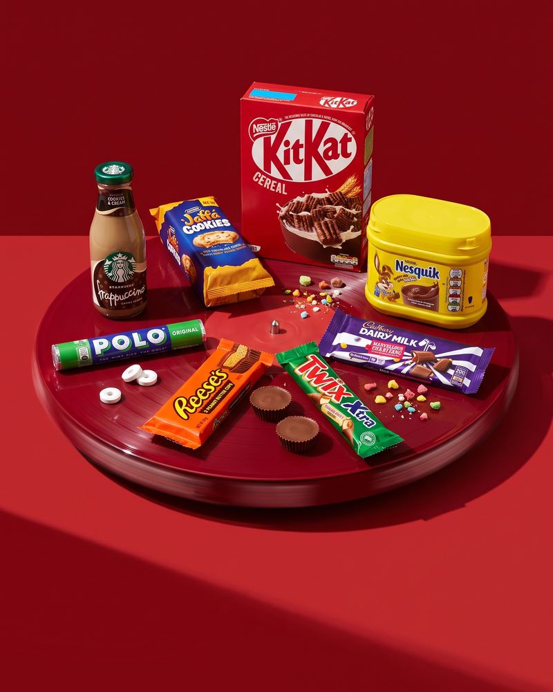 Starbucks Frappuccino, McVitie's Jaffa Cookies, a KitKat Cereal box, a Nesquik tub, Polo mints, Reese's Peanut Butter Cups, a Twix Xtra and a Cadbury Dairy Milk bar arranged on a glossy crimson turntable against a deep red backdrop.