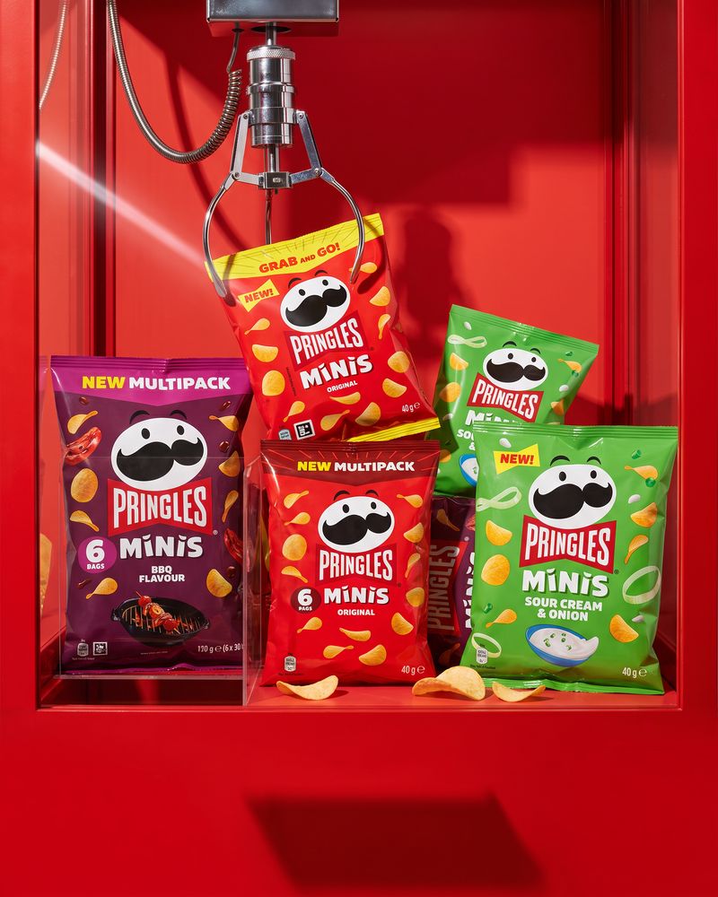 Pringles Minis bags and 6-pack multipacks inside a red arcade claw-machine cabinet, the chrome claw hanging above them.