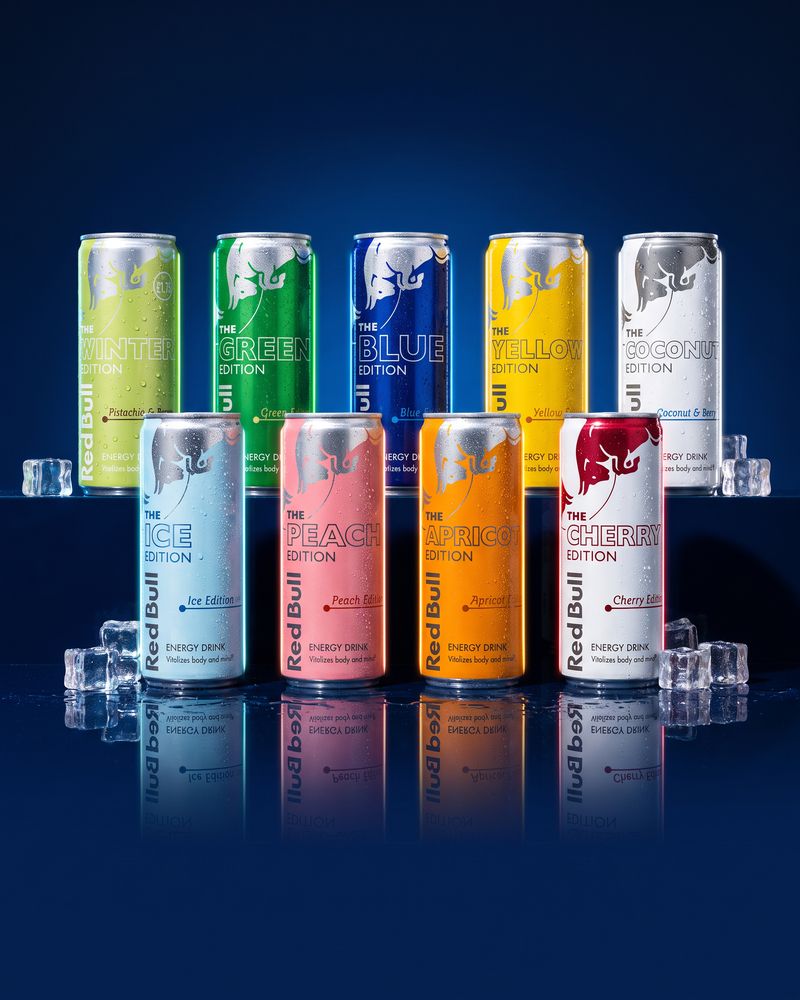 Nine Red Bull Edition 250ml cans standing in two rows on a wet dark-blue mirror floor with ice cubes, each can lit in its own edition colour — the Winter, Green, Blue, Yellow, Coconut, Ice, Peach, Apricot and Cherry Editions