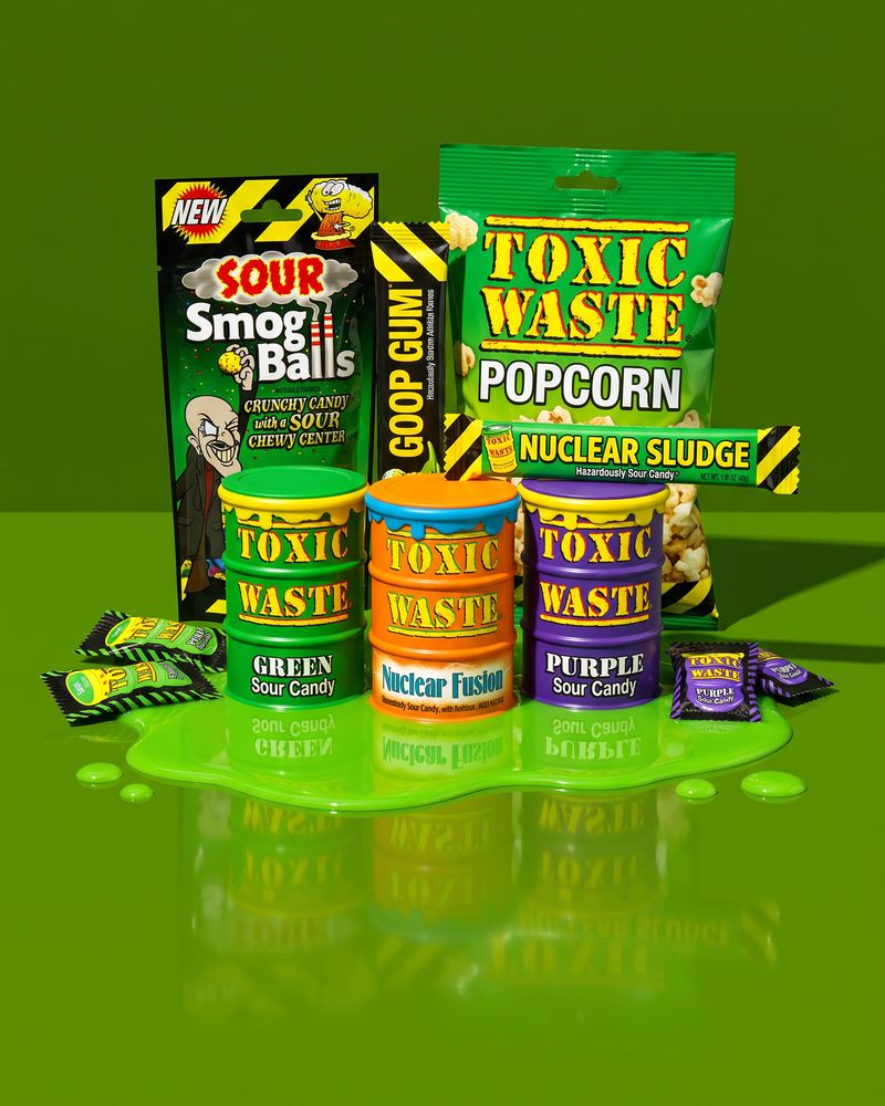Three Toxic Waste sour candy drums, a Sour Smog Balls pouch, Goop Gum, a popcorn bag and a Nuclear Sludge chew bar standing in a pool of green ooze on a glossy acid-green floor.
