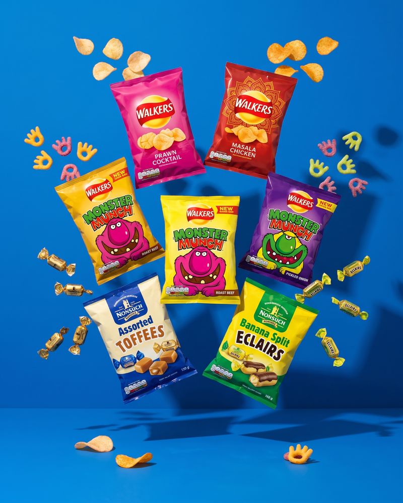 Plain foil crisp packets piled in a heap with golden crisps scattered in front, on a strong blue background.